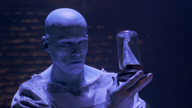 Screenshot of Arnold Schwarzenegger in Batman Forever. In his blue Mr. Freeze makeup, he holds a 5-inch-tall ice sculpture encased in a glass jar. The sculpture is a woman in a wedding dress and well-carved. Freeze wears striped a prison uniform and the background is a brick wall.